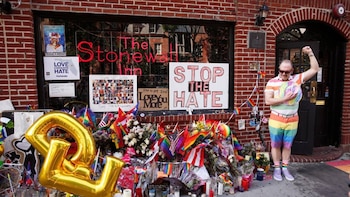 Fachada de ladrillo de Stonewall Inn con un gran memorial de flores, banderas arcoíris y velas. Un hombre con ropa de arcoíris y gafas de sol levanta el puño