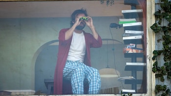 Hombre con barba y cabello desordenado, vestido con camiseta blanca, bata roja y pantalón de pijama a cuadros azules, mira con binoculares verdes por una ventana