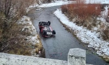 Automóvil rojo volcado en un arroyo. Nieve cubre las orillas y la vegetación. Parte de una estructura de concreto en primer plano