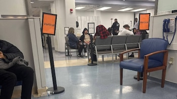 Sala de espera con personas sentadas en sillas grises y una silla individual azul en primer plano. Se ven letreros 'Vertical' y un teléfono de pared