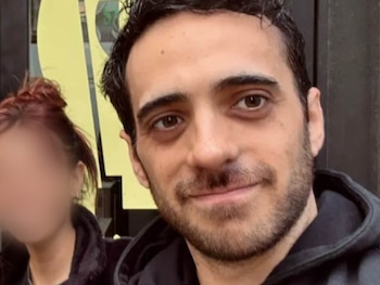 Primer plano de Alejandro Salazar, un hombre con cabello oscuro y barba, sonriendo levemente. Una persona con cabello rojizo y el rostro borroso aparece a su izquierda