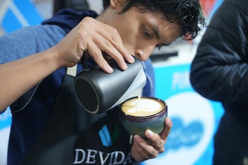 Un barista con delantal negro vierte leche de una jarra metálica oscura sobre una taza de cerámica con café, creando un diseño en la superficie
