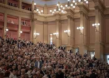 Salón de teatro clásico con columnas, lámparas de araña y balcones, ocupado por un público numeroso, hombres y mujeres vestidos de gala