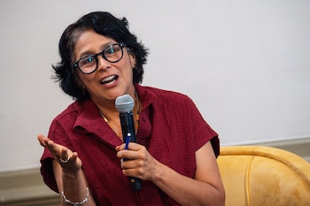 Mujer de cabello oscuro y gafas, vestida con camisa roja, habla en un micrófono con una mano extendida en gesto, sentada frente a fondo claro