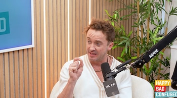 Tom Felton, de frente, gesticula mientras habla en un podcast. Viste un cárdigan blanco y beige, frente a un micrófono, con paredes de madera y plantas detrás