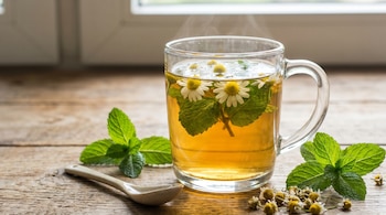 Una taza de vidrio transparente contiene una infusión humeante de manzanilla y menta, con flores y hojas frescas, sobre una superficie de madera.