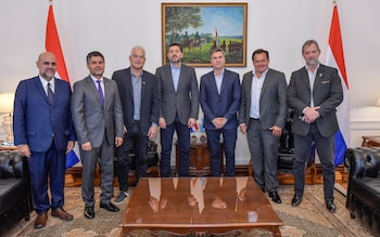 Siete hombres vestidos con traje de pie en una sala de reuniones con banderas de Paraguay y Argentina en el fondo y un cuadro colgado en la pared