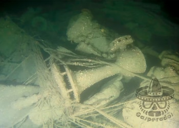 Imagen submarina de un naufragio, mostrando estructuras metálicas y escombros cubiertos de algas y sedimentos en aguas profundas y oscuras