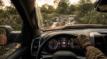 Vista desde el interior de una pickup roja con un conductor usando guantes en el volante, conduciendo por un camino rocoso y embarrado al atardecer.