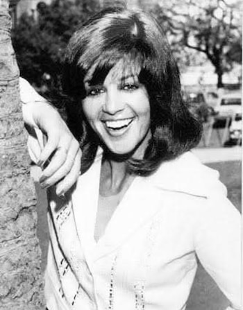 Fotografía en blanco y negro de una mujer sonriente con cabello oscuro y voluminoso, apoyada en el tronco de un árbol. Viste una camisa clara