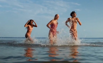 Dos mujeres y un hombre corren en aguas poco profundas de un cuerpo de agua. El movimiento genera salpicaduras. El cielo es azul