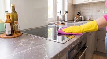 Una mano con guante amarillo limpia una encimera de cocina con un paño rosa. Se ven botellas, estufa, horno, fregadero, ventana y utensilios.