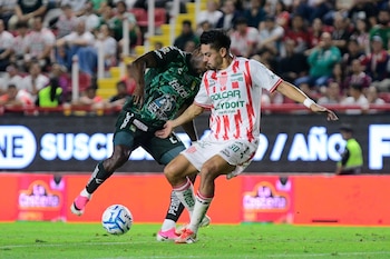 Dos futbolistas, uno con uniforme verde y otro con camiseta rojiblanca, disputan un balón en una cancha de césped con público en las gradas