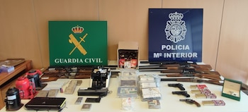 Una mesa blanca con varias armas de fuego, cajas de munición, paquetes rectangulares sellados, un reloj, herramientas y dos logos policiales al fondo