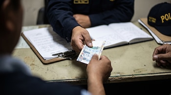Dos personas intercambian un billete de 100 soles peruanos sobre un escritorio con documentos, un libro abierto y una gorra de policía.