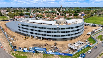 Vista aérea de un edificio curvo en construcción con grúas en la azotea, maquinaria pesada y vallas publicitarias en el frente