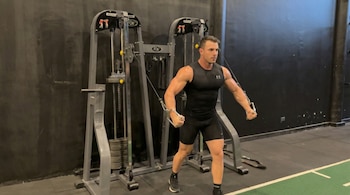 Un hombre musculoso en ropa deportiva negra realiza aperturas con polea en una máquina de gimnasio gris, con una pared oscura de fondo.