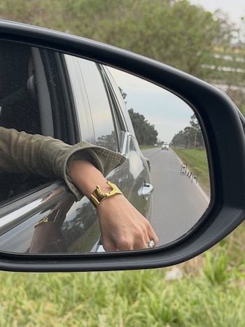 Espejo retrovisor lateral de un vehículo que refleja un brazo apoyado en la ventanilla con una pulsera dorada y un anillo
