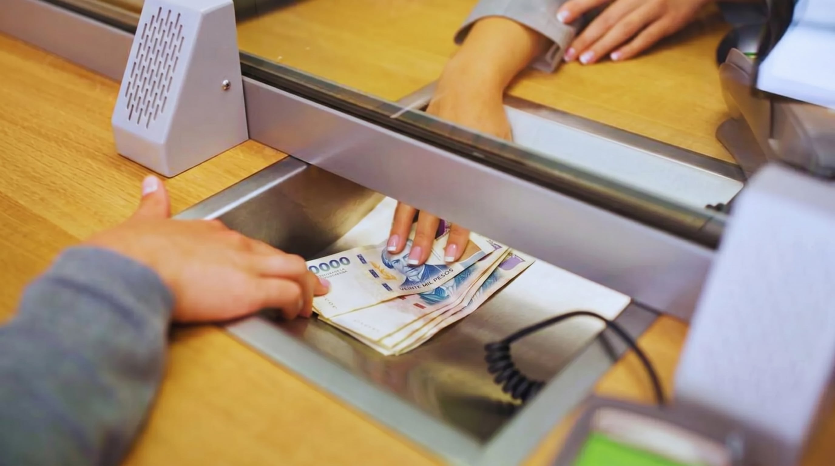 Dos manos interactúan en una ventanilla de servicio al cliente, con un fajo de billetes de pesos argentinos siendo entregado en la bandeja. (Imagen Ilustrativa Infobae)