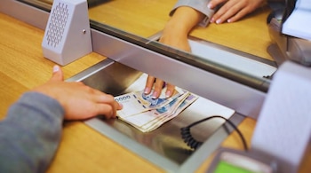 Primer plano de dos manos en un mostrador de madera, intercambiando un fajo de billetes de Pesos Argentinos a través de una bandeja metálica de ventanilla.