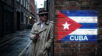 Un hombre con abrigo, sombrero, gafas de sol y teléfono celular junto a un muro con la bandera de Cuba y la palabra Cuba.