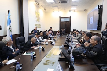 Un grupo de hombres sentados alrededor de una mesa larga en una sala de conferencias con la bandera de Guatemala, un logotipo del Congreso, monitores y botellas de agua