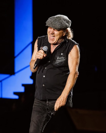 El vocalista Brian Johnson de AC/DC canta apasionadamente en un micrófono, vistiendo una gorra gris, camiseta negra sin mangas y jeans, en un escenario oscuro con luces azules