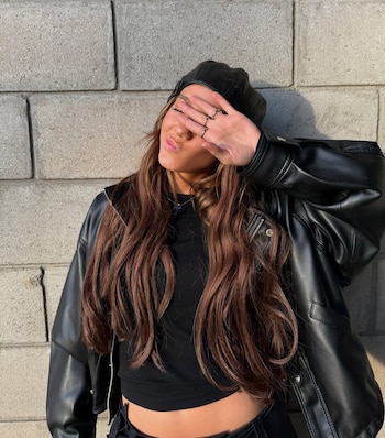 Mujer joven con cabello castaño largo, chaqueta de cuero negra y gorra negra, cubriendo su rostro con la mano. Fondo de pared de bloques grises
