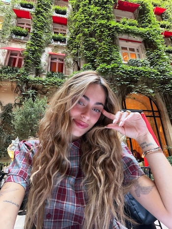 Retrato de una mujer joven con pelo rubio ondulado y camisa a cuadros, con brazaletes y tatuajes. Detrás, un edificio antiguo cubierto de hiedra y toldos rojos