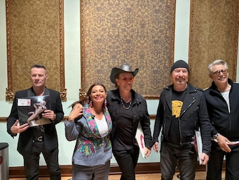 Cinco personas de pie en una sala con paredes decoradas: una mujer sonriente con blazer colorido junto a cuatro hombres, uno con sombrero y otro con gorro