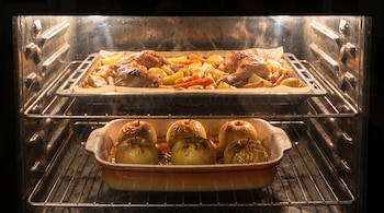 Interior de un horno iluminado con pollo, papas y zanahorias en la bandeja superior y seis manzanas en una fuente en la rejilla inferior.