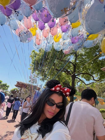 Charlotte Caniggia, con cabello oscuro, gafas de sol y orejas de Minnie Mouse con un lazo rojo, sonríe frente a globos de colores en un parque temático