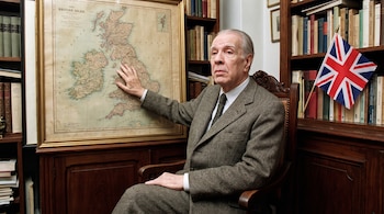 Un hombre sentado en un sillón de madera, con traje y corbata, su mano derecha sobre un mapa enmarcado de las Islas Británicas, y una bandera del Reino Unido.