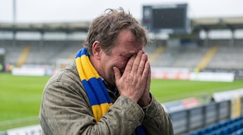 Primer plano de un hombre con una bufanda azul y amarilla, llorando y cubriéndose la cara con las manos en un estadio de fútbol vacío.
