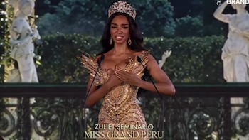 Una mujer con corona y vestido dorado tiene la mano en el pecho frente a dos micrófonos
