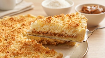 Una rebanada de tarta de coco tostado con relleno de dulce de leche es levantada con una espátula sobre un plato, con más tarta y cuencos en el fondo.