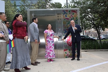 Varias personas asisten a la inauguración de un moderno baño público metálico con patrones florales coloridos; un hombre habla por un micrófono