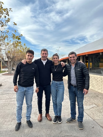 Cuatro personas (tres hombres y una mujer) de pie y sonriendo, con un brazo alrededor del hombro del otro, en un espacio exterior con pavimento, árboles y un edificio