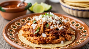 Plato de tinga de pollo con chipotle sobre tortilla, adornado con queso, cebolla y cilantro; al fondo, limones, salsa y una cesta de tortillas.