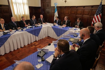 Un grupo de hombres con traje formal está sentado alrededor de una mesa en forma de U con las banderas de Guatemala y Estados Unidos