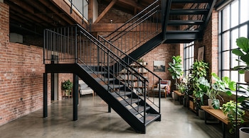 Escalera de acero negro, paredes de ladrillo visto, piso de hormigón, ventanas grandes, vigas de madera en el techo y plantas en macetas.