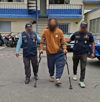 Un hombre con sudadera marrón y muletas camina asistido por dos agentes con uniforme de seguridad. Al fondo, un edificio azul y motos estacionadas