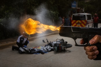 Mano que sostiene una pistola que expulsa fuego y humo, frente a un cuerpo cubierto en el suelo junto a una motocicleta.