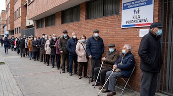 Una fila de adultos mayores con mascarillas quirúrgicas se extiende frente a un edificio de ladrillo. Dos hombres mayores se sientan en sillas plegables con bastones.