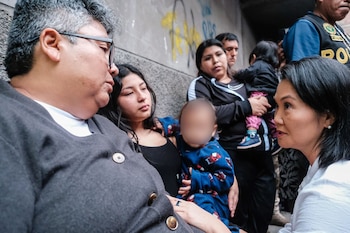 Una mujer con blusa blanca habla con otra mujer. Dos niños, un hombre y un oficial de policía están cerca. Fondo de pared con grafiti