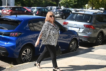Una mujer de cabello rojizo, gafas de sol, blusa con estampado blanco y negro, y pantalón oscuro camina en la vereda junto a autos estacionados