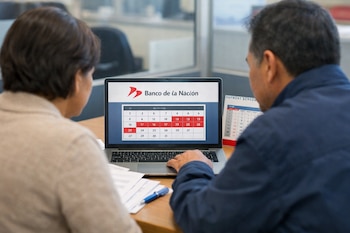 Dos personas de espaldas ven una computadora portátil con el logo del Banco de la Nación y un calendario de julio; hay documentos y un calendario físico en la mesa.