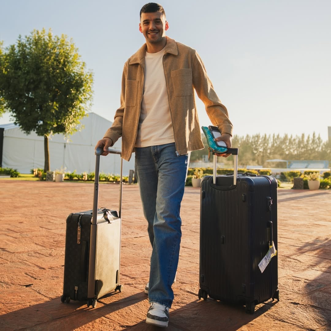 Gerónimo Rulli camina sonriente al aire libre, tirando de dos maletas en un día soleado en Ezeiza
