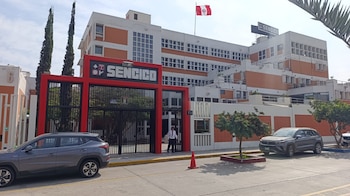 Vista exterior del edificio de Sencico en un día soleado, con el logo prominente en la entrada y en el techo, la bandera peruana ondeando y vehículos estacionados