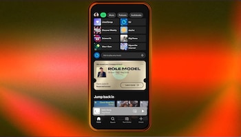 Un teléfono inteligente con la aplicación de Spotify abierta, mostrando una oferta para comprar dos entradas para el concierto de ROLE MODEL en un fondo degradado naranja y rojo